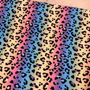 May include: A fabric featuring a rainbow leopard print. The background colours transition from yellow to pink, blue, and purple. Black leopard spots are scattered across the surface.