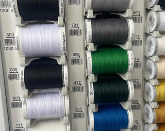 Puede incluir: Una exhibición de carretes de hilo Gütermann Sew-all en varios colores, incluyendo negro, blanco, gris, verde y azul. Los carretes están ordenados en un estante, con etiquetas que indican el tipo y la longitud del hilo. El texto "Sew-all Thread 500 m" es visible.