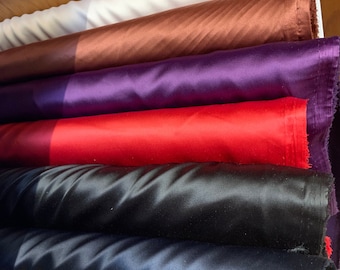 Puede incluir: Una selección de rollos de tela coloridos, incluyendo blanco, marrón, morado, rojo, negro y azul. Las telas tienen una textura brillante, similar al satén, y están cuidadosamente apiladas. La imagen muestra una variedad de colores y texturas, adecuadas para proyectos de costura y manualidades.