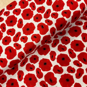 May include: A white fabric featuring a repeating pattern of red poppy flowers with black centres. The poppies vary in size and are arranged in different directions. The fabric is folded, revealing its texture and drape.