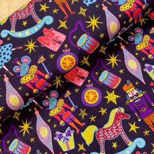 May include: A dark purple fabric featuring a repeating pattern of colourful holiday-themed illustrations including nutcrackers, mice in crowns, rocking horses, drums, gift boxes, and star shapes in shades of yellow, pink, blue, and red.