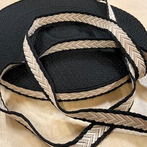 May include: Black woven ribbon with a beige and black chevron pattern. The ribbon is rolled up, with a length of the ribbon unrolled and looped over the roll. The ribbon is likely used for crafting or sewing projects.