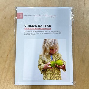 Könnte beinhalten: Ein Schnittmuster für einen Kinder-Kaftan, geeignet für Kinder im Alter von 1-2 und 2-3 Jahren. Die Verpackung zeigt ein Kind in einem gelben Blumen-Kaftan, das grüne Früchte hält. Der Text besagt, dass der Kaftan vor der Sonne schützt.