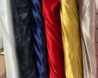 May include: A selection of rolled fabric bolts in various colors. The fabrics include white, black, navy blue, red, gold, and silver. The materials appear to be satin or a similar shiny textile, with visible folds and textures.