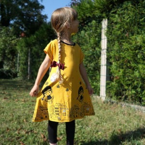 May include: A yellow dress with a black trim and a cityscape design. The dress features short sleeves and a flared skirt. The design includes cartoon figures and buildings. The dress is worn with black leggings.