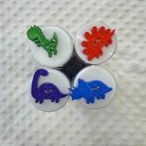 May include: Four colourful dinosaur buttons with a white background. The buttons are green, red, blue, and purple. Each button has a different dinosaur design.