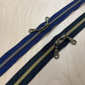 Könnte beinhalten: Zwei Reißverschlüsse mit antik-messingfarbenen Metallzähnen und -zippern. Ein Reißverschluss ist marineblau, der andere schwarz. Beide Reißverschlüsse liegen auf einer hellen Holzoberfläche.