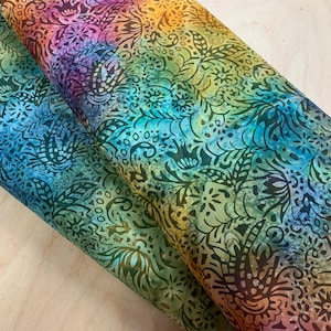 Puede incluir: Tela enrollada con un vibrante diseño batik multicolor. La tela presenta una transición de colores de azul a verde, amarillo, naranja y morado, con un detallado patrón floral y de hojas en marrón oscuro. El material parece suave.