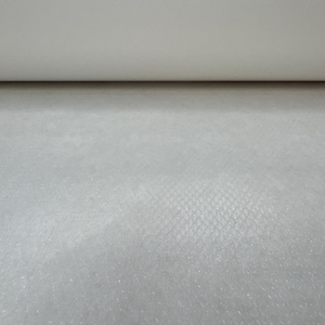 May include: A close-up of a roll of white fabric. The fabric has a textured surface with a subtle pattern. The top portion of the image shows the rolled edge of the fabric, while the rest displays the textured material.