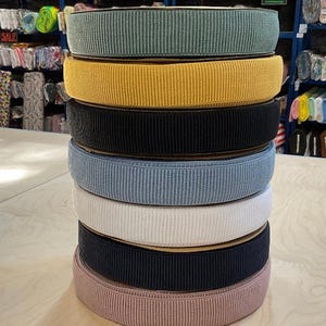 Puede incluir: Una pila de ocho gomas elásticas de diferentes colores. Las gomas son todas acanaladas y tienen una superficie ligeramente texturizada. Los colores son verde azulado, amarillo, negro, azul claro, blanco, negro y rosa.