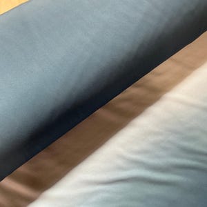 May include: A roll of fabric featuring a smooth, solid dark teal surface that transitions into a lighter, gradient ombre effect, fading from a deep blue-green hue into a soft, pale white tone across the length of the material.