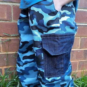May include: Blue camouflage pants with a dark blue denim pocket. The pants feature a pattern of light and dark blue shapes. The person is standing in front of a brick wall and green grass.