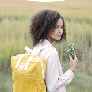 May include: A person wearing a pink shirt and blue jeans is standing in a field of tall grass. They are holding a bouquet of small white flowers in one hand and wearing a yellow canvas backpack with white straps and a front pocket. The backpack has the text "MAKING BACKPACK" and "a simple backpack with interior zip pocket and front pocket" above the image. The text "noodlehead" is at the bottom of the image.