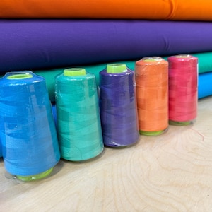 May include: Six spools of thread in various colours, including blue, green, orange, and pink. The spools are arranged in a row on a wooden surface.