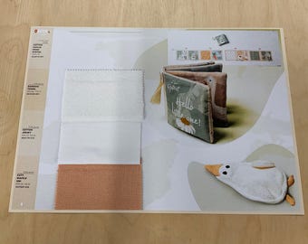 Peut inclure: Présentation sur le thème des bébés avec un livre souple portant l'inscription « Hello little one ! » et un jouet en forme d'oie en peluche. L'affichage comprend également des échantillons de tissu blancs et pêche.