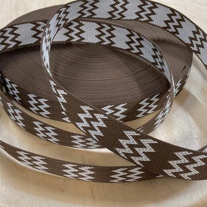 Op de afbeelding: Een rol bruin lint met een wit en lichtgrijs zigzagpatroon. Het lint is strak opgerold, met een deel afgerold om het ontwerp te laten zien. Dit lint is geschikt voor diverse knutsel- en decoratieprojecten.