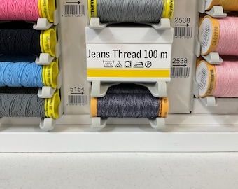 Puede incluir: Primer plano de carretes de hilo en varios colores, incluyendo rosa, negro, azul y gris. El texto "Jeans Thread 100 m" es visible en una etiqueta. Los carretes están organizados en una vitrina, con códigos de barras y otro texto.