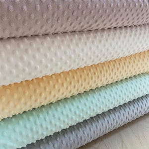 Könnte beinhalten: Ein Stapel weicher, strukturierter Stoffe in verschiedenen Pastellfarben. Die Stoffe haben ein erhabenes Punktmuster. Zu den Farben gehören Hellviolett, Weiß, Gelb, Mintgrün und Grau. Der Stoff scheint ein Plüschmaterial zu sein.