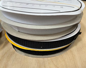 May include: A stack of spools of white and black hook and loop tape. The rolls are stacked on top of each other, with a yellow edge visible on one spool. The top spool has text printed on it.