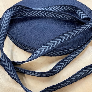May include: A roll of navy blue woven ribbon with a repeating grey chevron pattern along the edges. The ribbon is neatly coiled, with a length of the patterned ribbon extending outwards. The ribbon is likely used for crafting or sewing projects.
