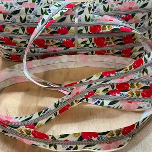 Könnte beinhalten: Eine Rolle Reißverschlussband mit floralem Muster mit weißem Hintergrund und roten, rosa und grünen Blumenmustern. Das Reißverschlussband ist silberfarben. Das Band liegt auf einer hellbraunen Holzoberfläche.