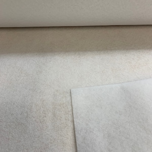 Könnte beinhalten: Eine Rolle cremefarbenen, strukturierten Stoffes, möglicherweise Filz oder ein ähnliches Material, ist teilweise abgerollt und zeigt eine gefaltete Kante. Der Stoff hat ein weiches, leicht gesprenkeltes Aussehen mit subtilen Farbvariationen.