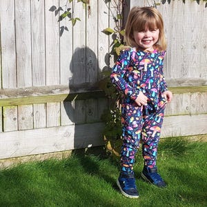 May include: A young person wearing a blue and black long-sleeved jumpsuit with a colourful mushroom print. The jumpsuit has a crew neck and long sleeves. The person is standing in front of a wooden fence and smiling.