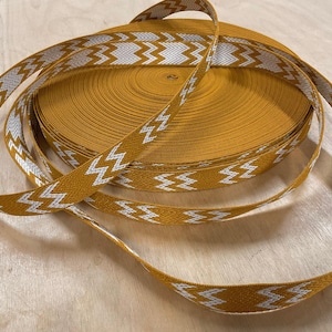 Peut inclure: Un rouleau de ruban jaune moutarde avec un motif en zigzag blanc. Le ruban est enroulé, avec une longueur du ruban à motifs déroulée et posée dessus. Le ruban est adapté aux travaux manuels et à la décoration.