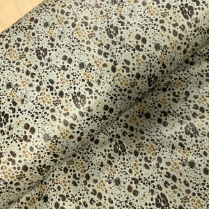 May include: A roll of fabric featuring a cream-coloured base with a speckled pattern in shades of brown and tan. The fabric has a textured appearance, with the pattern resembling animal print.