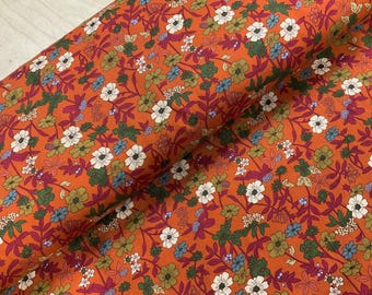 May include: A roll of fabric with a vibrant floral pattern. The fabric features a burnt orange background with a repeating design of white, green, blue, and burgundy flowers and leaves. The fabric is likely cotton or a similar material.