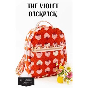 May include: A small, round backpack with a red background and a pattern of pink strawberries. The backpack has a zipper pocket, a handle, and adjustable straps. The text "THE VIOLET BACKPACK" is at the top of the image. The backpack is approximately 25 cm in height.