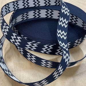 Könnte beinhalten: Eine Rolle marineblaues Band mit weißem Zickzackmuster. Ein Teil des Bandes ist abgerollt und über die Oberseite gelegt. Das Band eignet sich für verschiedene Bastelprojekte, wie Nähen oder Geschenkverpackung. Messung in Zentimetern.