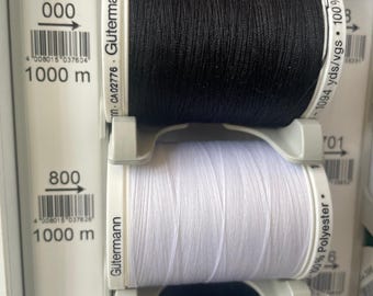 Puede incluir: Tres carretes de hilo, uno negro, uno blanco y uno negro. Cada carrete está etiquetado con el nombre de la marca "Gütermann" y la longitud del hilo en metros. Los carretes negros están etiquetados con "000" y el carrete blanco está etiquetado con "800".