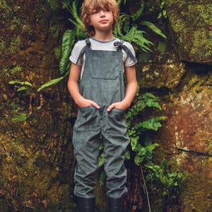 May include: A young person wearing a dark green overall with a white t-shirt and black wellies. They are standing in front of a mossy rock wall with green ferns.