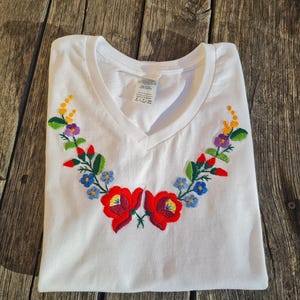 Può includere: Maglietta bianca a collo a V con un ricamo floreale colorato sul davanti. Il disegno presenta fiori rossi, gialli, blu, verdi e viola.