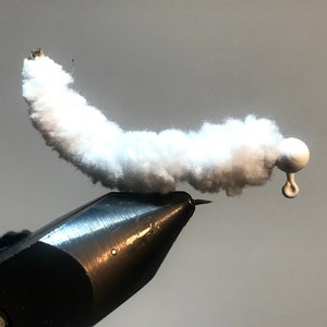 Op de afbeelding: Een wit vliegenbindmateriaal is vastgemaakt aan een zwart vliegenbindgereedschap. Het materiaal is pluizig en lijkt op een rups. Een witte kraal is bevestigd aan het uiteinde van het materiaal.