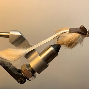 May include: A fly fishing lure with a tan body and a dark brown head. The lure is attached to a silver hook and held in a silver vise. The lure has a white strap and is designed to mimic an insect.