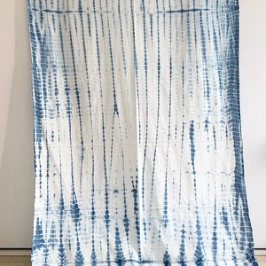 May include: White textile with a blue tie-dye pattern. The design features vertical lines and dotted patterns in varying widths, creating a textured appearance. The fabric is draped, showcasing the full length of the textile.