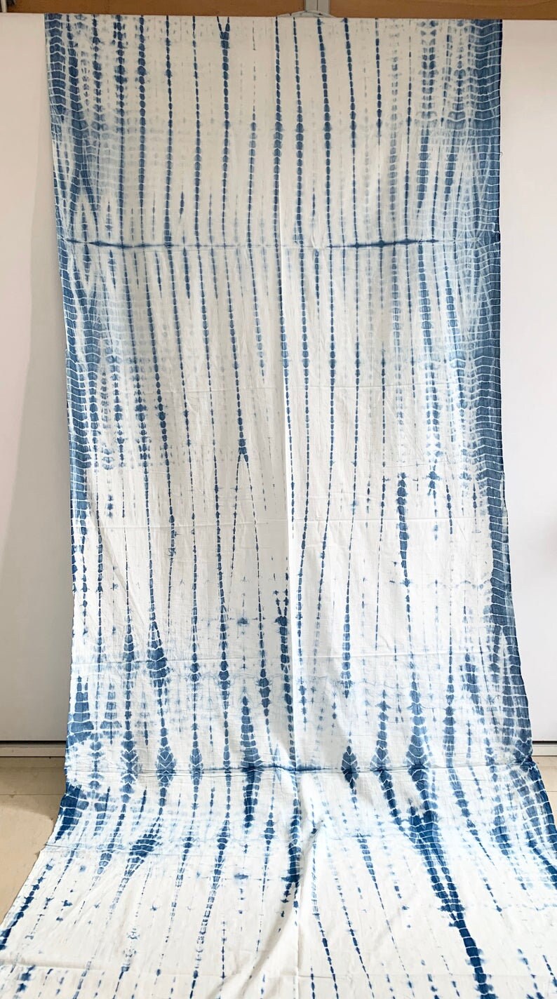 May include: White textile with a blue tie-dye pattern. The design features vertical lines and dotted patterns in varying widths, creating a textured appearance. The fabric is draped, showcasing the full length of the textile.