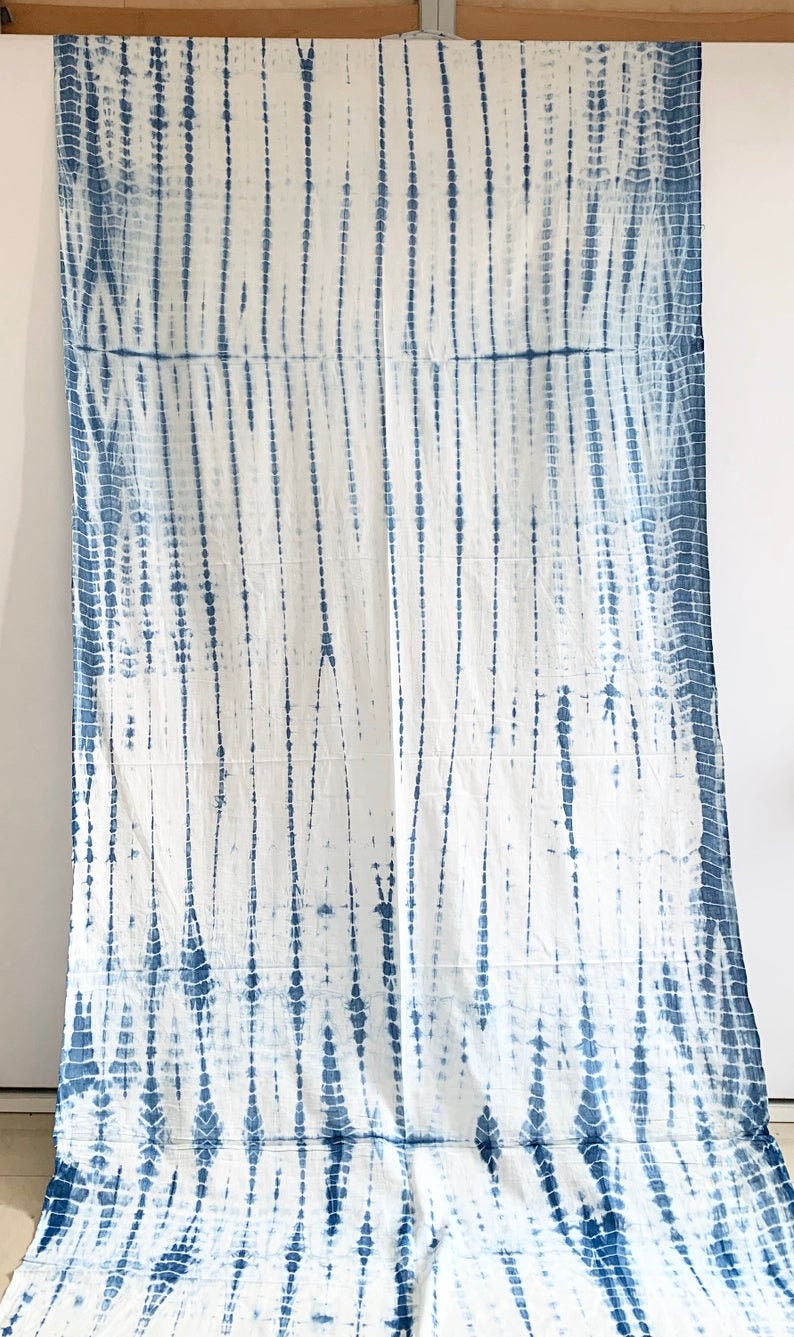 May include: White fabric with a blue tie-dye pattern. The design features vertical lines of varying widths and dotted patterns, creating a textured appearance. The fabric is hanging against a white wall.