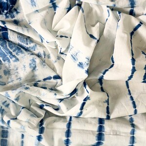 May include: White fabric with a blue tie-dye pattern. The fabric has a crinkled texture, with vertical and horizontal blue lines and shapes against a white background. The design gives the fabric a handcrafted appearance.