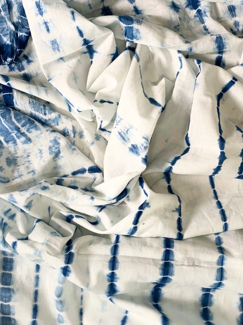 May include: White fabric with a blue tie-dye pattern. The fabric has a crinkled texture, with vertical and horizontal blue lines and shapes against a white background. The design gives the fabric a handcrafted appearance.