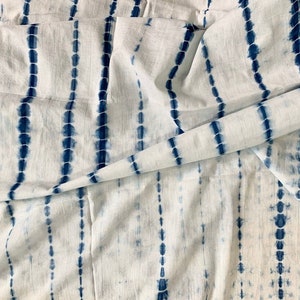 May include: White fabric with a blue tie-dye pattern. The design features vertical stripes of blue dots and dashes, creating a textured effect. The fabric is folded, showing the pattern's continuity.