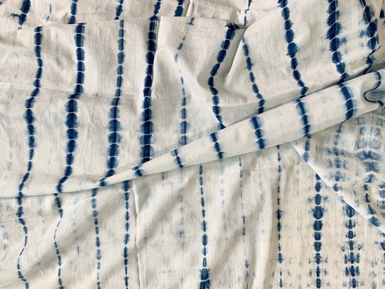 May include: White fabric with a blue tie-dye pattern. The design features vertical stripes of blue dots and dashes, creating a textured effect. The fabric is folded, showing the pattern's continuity.
