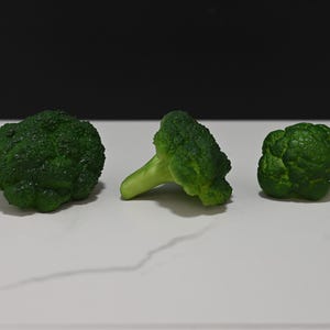 May include: Three green broccoli florets on a white marble surface. The florets are arranged in a row, with the middle floret facing the viewer.