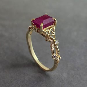 May include: A gold ring with a rectangular, deep red gemstone. The ring has an intricate, Celtic-inspired design with a knot detail and small, clear accent stones. The band has a textured, organic appearance.
