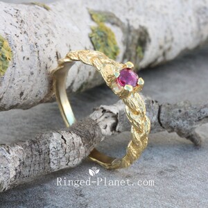 May include: A gold ring featuring a textured, plaited band and a round, magenta gemstone. The ring is set against a natural backdrop, emphasising its organic design. The website "Ringed-Planet.com" is visible at the bottom.