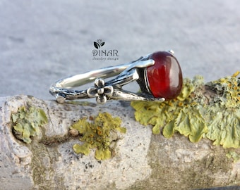 May include: A silver ring with a red gemstone set in a branch-like design. The ring has a small flower detail on the side.