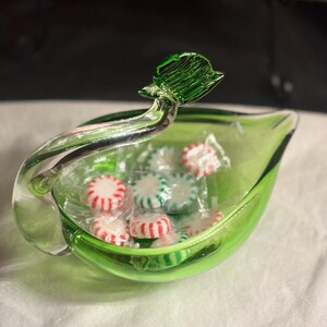 May include: A bright green glass bowl shaped like a leaf with a clear glass handle extending upward. The bowl contains several small, round, individually wrapped peppermint candies featuring red and white stripes inside clear plastic wrappers.