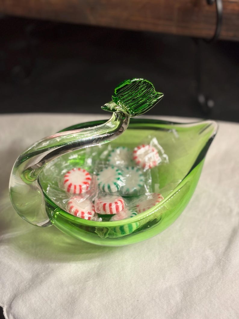 May include: A bright green glass bowl shaped like a leaf with a clear glass handle extending upward. The bowl contains several small, round, individually wrapped peppermint candies featuring red and white stripes inside clear plastic wrappers.